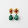 Pair of earrings with gold clasps, red stones, and green teardrop-shaped beads on a beige fabric background.