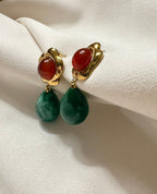 Pair of earrings with gold clasps, red stones, and green teardrop-shaped beads on a beige fabric background.