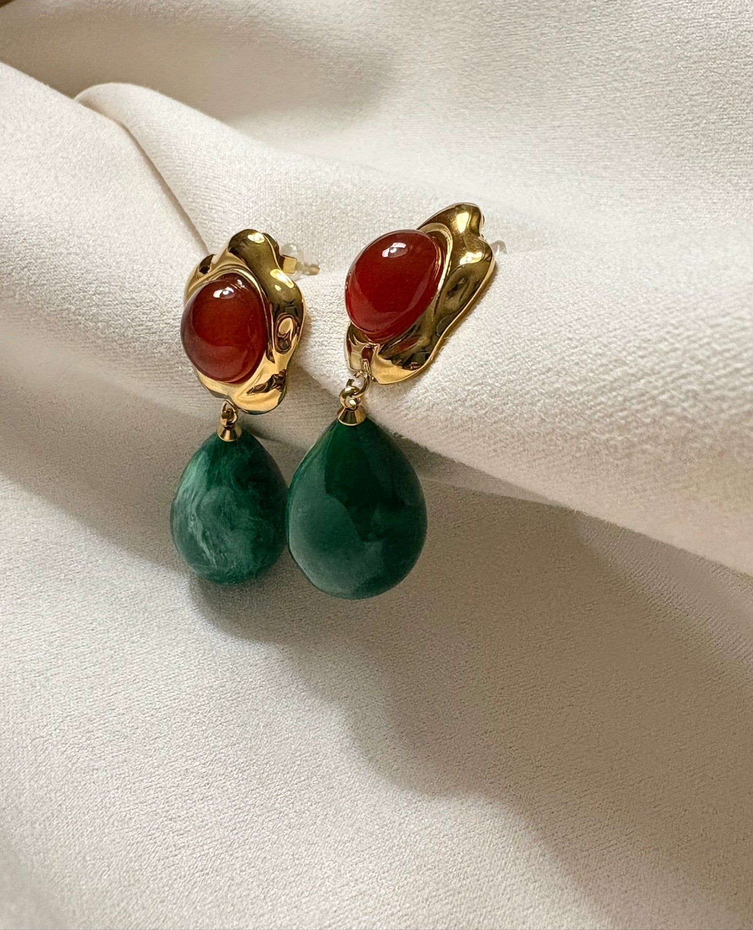 Pair of earrings with gold clasps, red stones, and green teardrop-shaped beads on a beige fabric background.